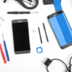 Organized workspace with precision screwdrivers, plastic spudgers, tweezers, anti-static wrist strap, and heat gun laid out on clean white surface next to open Samsung smartphone