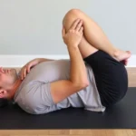 Person performing knee-to-chest stretch on yoga mat, lying on back with one knee drawn toward chest, demonstrating proper lower back mobility exercise technique