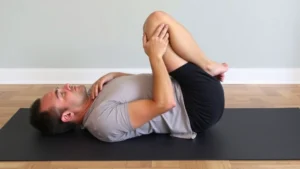 Person performing knee-to-chest stretch on yoga mat, lying on back with one knee drawn toward chest, demonstrating proper lower back mobility exercise technique