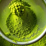 Close-up of vibrant bright green matcha powder being sifted through fine mesh strainer into ceramic bowl, showing fine powder particles in detail