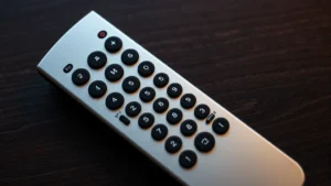Close-up overhead view of a silver universal remote control with illuminated buttons on dark wooden table, showing setup and mode buttons clearly