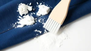 Close-up of white deodorant residue on dark blue fabric being gently brushed away with a soft toothbrush, showing white powder falling onto white surface, natural lighting
