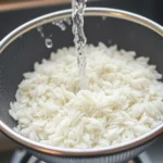 Close-up of fine-mesh strainer holding white rice under running cool water, rice grains visible through clear plastic strainer, kitchen sink background, daylight from window