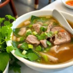 A steaming bowl of authentic pho with fresh herbs, lime, and beef in clear broth, served in a traditional Vietnamese restaurant setting with chopsticks and spoon on white tablecloth